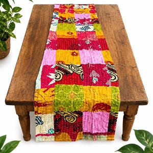Könnte beinhalten: Ein rechteckiger Tischläufer mit einem Patchwork-Design aus verschiedenen Stoffquadraten in leuchtenden Rosa-, Gelb-, Rot- und Grüntönen. Das Textil zeigt komplizierte Muster wie Wirbel, florale Motive und geometrische Formen mit sichtbaren weißen Nähten.