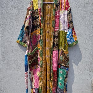 May include: A long-sleeved robe featuring a patchwork design made from various colorful fabric scraps in shades of yellow, pink, green, blue, and red. The garment includes a mustard yellow inner lining, two front pockets, and a matching fabric tie belt.