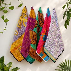 May include: Five rectangular quilted fabric pieces hanging vertically, each featuring distinct patterns and colors including yellow with purple floral motifs, magenta with geometric shapes, green with brown leaf prints, pink with blue accents, and white with purple flowers.