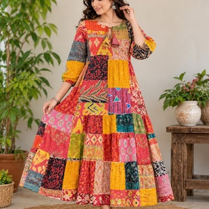 May include: A long-sleeved, ankle-length dress featuring a vibrant patchwork design with various colorful patterns in shades of red, yellow, orange, and blue. The dress has a round neckline with a tie detail and a tiered, flared skirt.