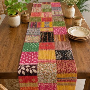May include: A rectangular table runner featuring a patchwork design composed of various fabric squares in vibrant colors including red, yellow, pink, green, and black. The runner displays diverse floral, geometric, and abstract patterns with visible stitching.