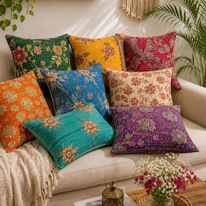 Puede incluir: Una colección de ocho cojines cuadrados con intrincadas costuras estilo kantha y vibrantes patrones florales en verde, amarillo, rojo, naranja, azul, crema, turquesa y morado, dispuestos juntos sobre un sofá de color claro.