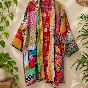 Könnte beinhalten: Eine langärmelige, vorne offene Jacke mit einem Patchwork-Design aus verschiedenen bunten Stoffen mit komplexen Mustern. Das Kleidungsstück verfügt über zwei Vordertaschen mit schwarzer Einfassung und sichtbaren weißen Nähten auf der Steppung.