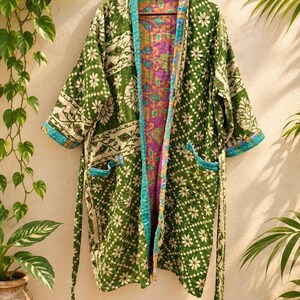 May include: A long-sleeved open-front robe featuring a green exterior with a white floral and geometric pattern. The garment includes two front pockets, a matching tie belt, and a contrasting interior lining with a purple and gold paisley print.