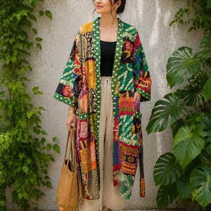 Könnte beinhalten: Eine lange, vorne offene Kimono-Jacke mit einem lebendigen Patchwork-Design aus verschiedenen Blumen- und geometrischen Mustern in Grün-, Rot-, Gelb- und Schwarztönen. Das Kleidungsstück hat weite Ärmel, zwei Vordertaschen und einen dunkelgrünen Saum.