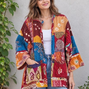 May include: A long-sleeved, open-front jacket featuring a patchwork design with various colourful fabric squares in shades of red, blue, yellow, and orange. The garment includes two front pockets and visible white stitching throughout the fabric.