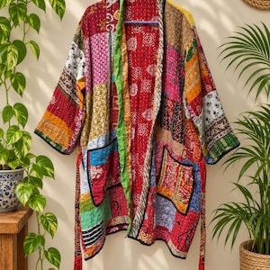 May include: A long-sleeved kimono-style jacket featuring a patchwork design of various colourful fabrics with intricate patterns. The garment includes two front pockets, a tie belt, and visible white stitching throughout the quilted material.