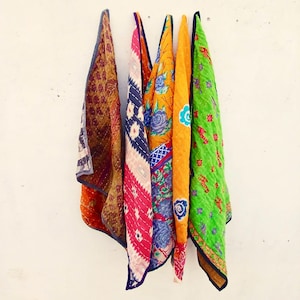 May include: A display of vibrant, patterned textiles hanging against a white wall. The fabrics showcase floral and geometric designs in shades of orange, pink, blue, and green. The edges are trimmed with contrasting colours.