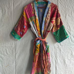 Könnte beinhalten: Bunter Patchwork-Bademantel mit Bindegürtel, an einem Holzbügel hängend. Der Bademantel weist eine Vielzahl von Mustern und Farben auf, darunter Rosa, Rot, Gelb, Grün und Blau. Die Ärmel sind dreiviertellang.