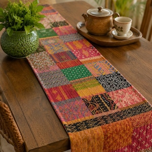 May include: A rectangular table runner featuring a patchwork design composed of various fabric squares in vibrant colors including red, orange, green, and black. The runner has visible quilted stitching throughout and displays a variety of patterns.
