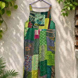 May include: A sleeveless jumpsuit featuring a patchwork design of various green, yellow, and multi-colored fabric scraps. The garment is constructed with visible white running stitch quilting throughout and hangs from a light purple plastic hanger.