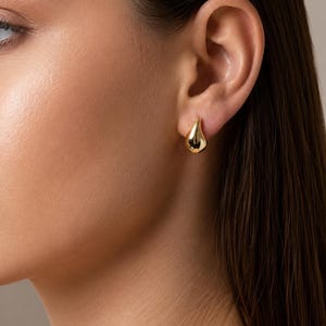 May include: A close-up of an ear with a gold teardrop earring. The earring reflects light, with a smooth surface. The skin is fair, and the hair is dark brown. The earring is a stylish accessory.