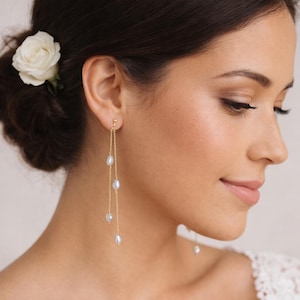 Peut inclure: Une paire de boucles d'oreilles pendantes dorées composées de deux chaînes fines suspendues à un clou. Chaque chaîne est ornée de petites perles blanches en forme de goutte d'eau espacées, créant un effet de superposition pendant.