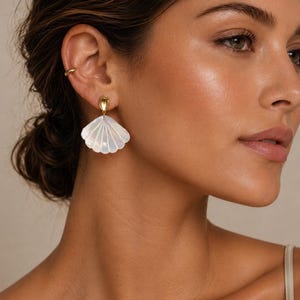 Peut inclure: Gros plan sur une oreille portant un ear cuff doré et une boucle d'oreille blanche en forme de coquillage aux détails irisés. La boucle d'oreille a un clou doré. La personne a les cheveux et la peau bruns.