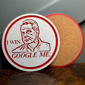 May include: A round white coaster with a red border and the text "I WIN. GOOGLE ME." features a red illustration of a man's face. A second coaster with a cork surface and red border is next to it.