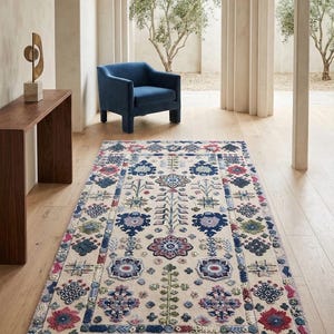 May include: A rectangular area rug with a cream-colored background and a detailed floral pattern in shades of blue, red, and green. The rug is placed on a wooden floor, with a blue armchair and a wooden table in the background.