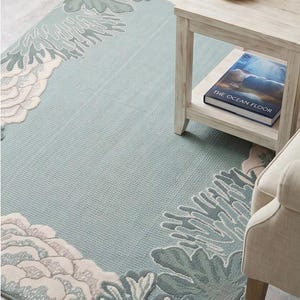 Peut inclure: Un tapis bleu clair rectangulaire avec un motif de récif corallien blanc, vert clair et gris. Le tapis a une surface texturée. Une petite table d'appoint en bois avec un livre intitulé "The Ocean Floor" est à côté du tapis.