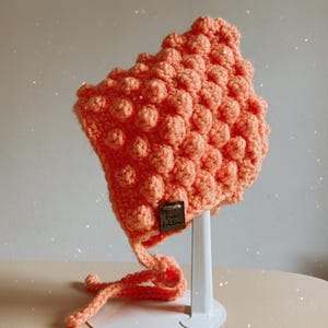 May include: A coral-colored, handmade crochet bonnet with a textured, bobble stitch design. The bonnet has ear flaps and ties, and a small tag is sewn on the front. The bonnet is displayed on a white stand against a neutral background.