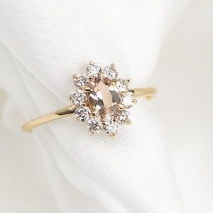 May include: A gold ring featuring a central oval-shaped peach-colored gemstone surrounded by smaller, clear gemstones. The ring's band is thin and gold, set against a white background. The ring is a piece of jewelry.