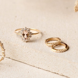May include: A collection of gold rings. One ring features a peach-colored oval gemstone surrounded by small, clear stones. Two small hoop earrings are also visible. The rings are set on a neutral-toned surface.