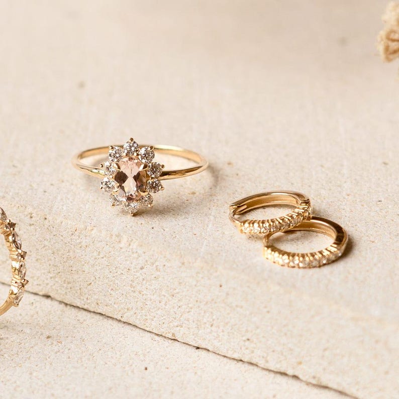 May include: A collection of gold rings. One ring features a peach-colored oval gemstone surrounded by small, clear stones. Two small hoop earrings are also visible. The rings are set on a neutral-toned surface.