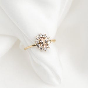 May include: A gold ring featuring a central oval-shaped peach-colored gemstone surrounded by smaller, clear gemstones. The ring's band is thin and gold, set against a white background. The ring is a piece of jewelry.