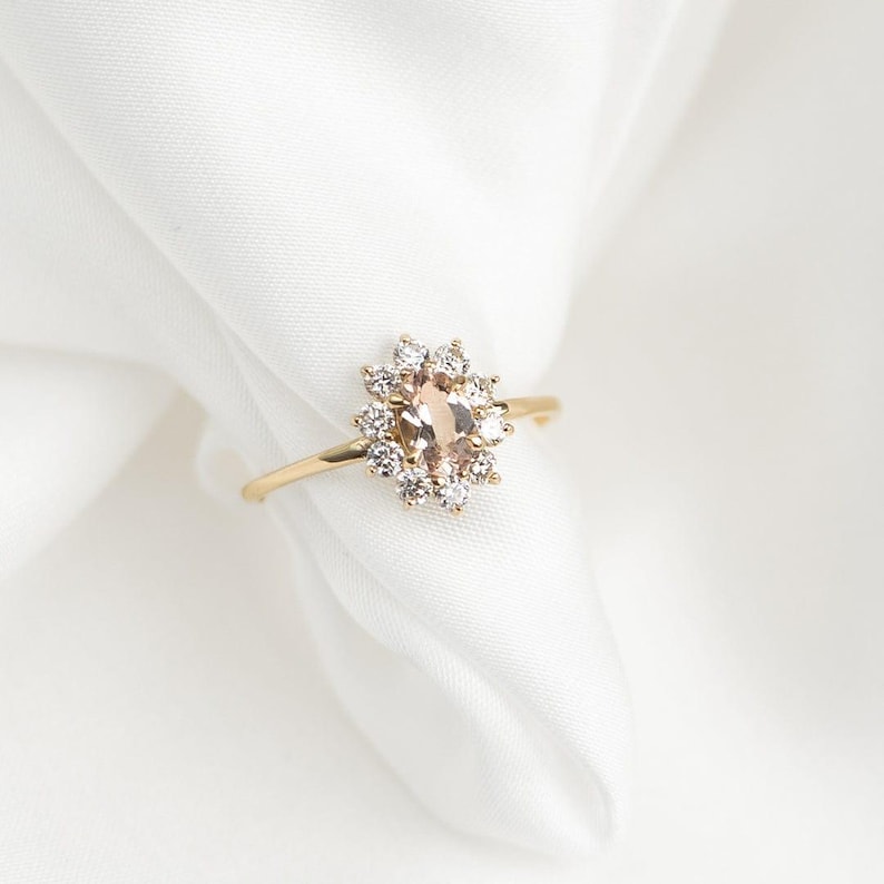 May include: A gold ring featuring a central oval-shaped peach-colored gemstone surrounded by smaller, clear gemstones. The ring's band is thin and gold, set against a white background. The ring is a piece of jewelry.