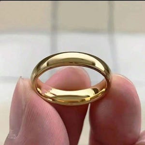 Peut inclure: Une bague dorée à la surface lisse et polie. La bague est de conception simple, reflétant la lumière. La bague est tenue entre deux doigts. Le diamètre de la bague est d'environ 2 cm.