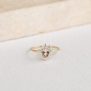May include: A gold ring featuring a central oval-cut peach-colored gemstone, surrounded by a halo of smaller, clear gemstones. The ring's band is thin and gold, creating a delicate and elegant design. The ring is set against a white background.