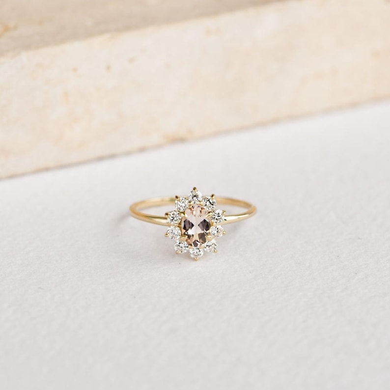 May include: A gold ring featuring a central oval-cut peach-colored gemstone, surrounded by a halo of smaller, clear gemstones. The ring's band is thin and gold, creating a delicate and elegant design. The ring is set against a white background.