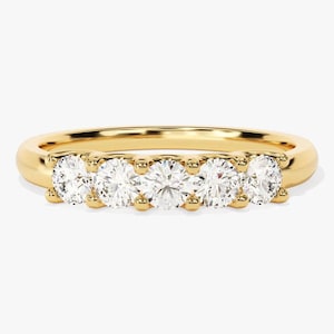 May include: A gold band ring featuring five round, clear gemstones. The gemstones are evenly spaced across the top of the ring, set in individual prongs. The ring has a polished, smooth finish and is set against a white background.