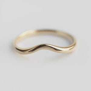 May include: A curved gold ring with a smooth, polished surface. The ring has a gentle wave design, creating a unique and elegant look. The ring is likely made of gold and is designed to be worn on a finger. The ring's design is simple yet sophisticated.