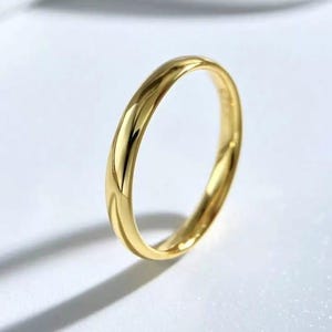 Könnte beinhalten: Nahaufnahme eines glänzenden Goldrings. Die glatte Oberfläche des Rings reflektiert das Licht und erzeugt Glanzlichter und Schatten. Der Ring liegt vor einem weißen Hintergrund, mit einem Schatten nach links.