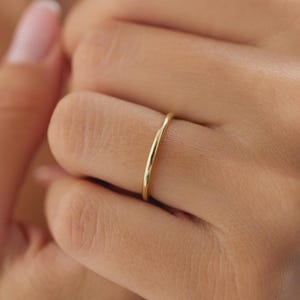 May include: A slender, gold-coloured ring is worn on a finger. The ring has a simple, minimalist design and a smooth, polished surface. The ring appears to be made of a precious metal, such as gold.