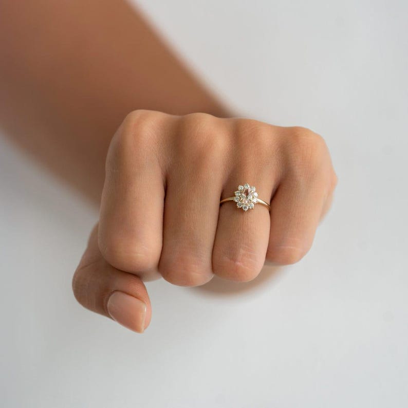May include: A gold ring featuring a cluster of small, round, clear gemstones arranged in a floral pattern. The ring is set on a thin gold band. The ring is on a hand with a neutral skin tone.
