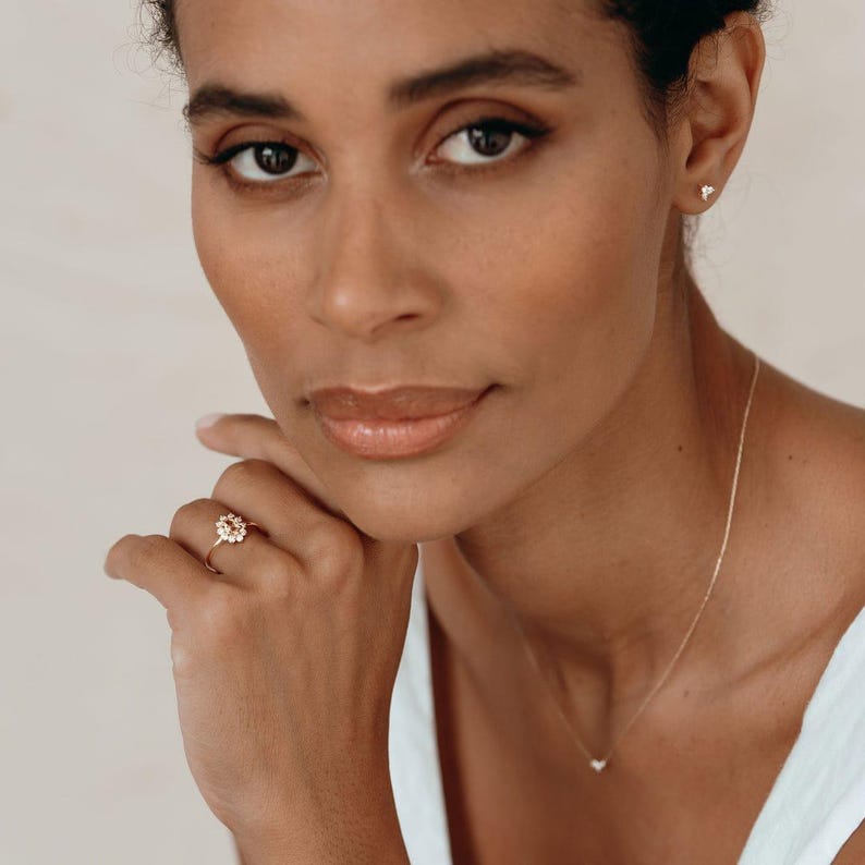 May include: A gold ring with a floral design featuring small, round gemstones. The ring is worn on a finger. A delicate gold necklace with a small pendant is also visible. The model is wearing a white top.
