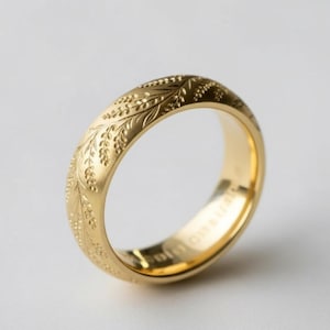 Puede incluir: Un anillo de oro con un patrón floral grabado. El anillo tiene un acabado mate y un interior pulido. El diseño presenta hojas detalladas y pequeñas formas de bayas. El anillo es un estilo de banda clásico.