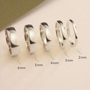 Puede incluir: Se muestran cinco anillos plateados de diferentes anchos. Los anillos miden 6 mm, 5 mm, 4 mm, 3 mm y 2 mm. Los anillos están muy pulidos y reflejan la luz, mostrando sus superficies lisas y redondeadas.