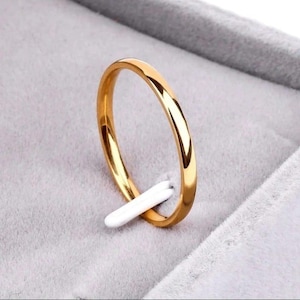 May include: A gold ring with a polished finish. The ring is presented on a white ring holder within a grey jewellery box. The ring is a simple band, reflecting light. The ring is likely made of gold.