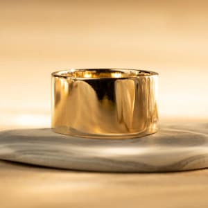 May include: A wide, gold-colored ring with a polished, reflective surface. The ring is cylindrical in shape and sits on a gray and white marbled surface. The background is a warm, golden hue.