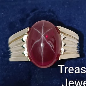 May include: A silver ring with a large, oval, deep red gemstone displaying a star effect. The ring is set on a dark blue velvet surface, with the text "Treasured Jeweller" visible.