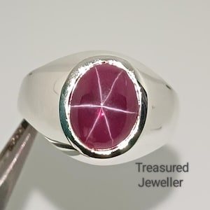 Peut inclure: Bague en argent avec une pierre précieuse ovale rouge foncé. La pierre présente un motif en forme d'étoile au centre. La bague est tenue par une pince. Le texte "Treasured Jeweller" est visible.