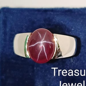 Peut inclure: Bague en argent ornée d'un grand rubis étoilé ovale. Le rubis est d'un rouge profond avec un motif en étoile à six rayons. La bague est posée sur un tissu de velours bleu foncé. Le texte "Treasured Jeweller" est visible.