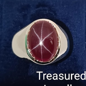 Peut inclure: Bague en argent avec une pierre précieuse ovale rouge foncé, ornée d'un motif en étoile à six rayons. La bague est posée sur une surface en velours bleu foncé. Le texte "Treasured Jeweller" est visible.