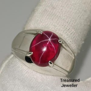 May include: A silver ring featuring a large, oval, deep red gemstone with a star-like pattern. The ring has a classic, rectangular design with a polished finish. The text "Treasured Jeweller" is visible.
