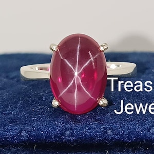 Peut inclure: Bague en argent ornée d'une grosse pierre rouge foncé ovale avec un motif en étoile à six branches. La bague est posée sur une surface bleue, avec le texte "Treasured Jeweller" visible.