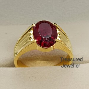 Puede incluir: Un anillo de oro con una gran gema roja oscura de forma ovalada. El anillo tiene una banda texturizada y se muestra en una caja de joyería verde. Las palabras "Treasured Jeweller" son visibles en la caja.
