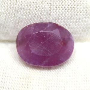 May include: A faceted, oval-shaped ruby gemstone. The gem is a deep, saturated pinkish-red color, with visible internal inclusions. The stone is set against a white, textured fabric background, highlighting its color and clarity. This gemstone is suitable for jewelry making.