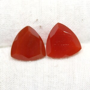 May include: Two triangular, faceted gemstones with a vibrant, reddish-orange hue. The gems are displayed on a white, textured surface, showcasing their polished surfaces and geometric shapes. These gemstones are likely for jewelry making.