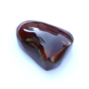May include: A polished, dark brown gemstone with a smooth, rounded shape. The surface reflects light, creating highlights and a glossy appearance. The stone is set against a white background.